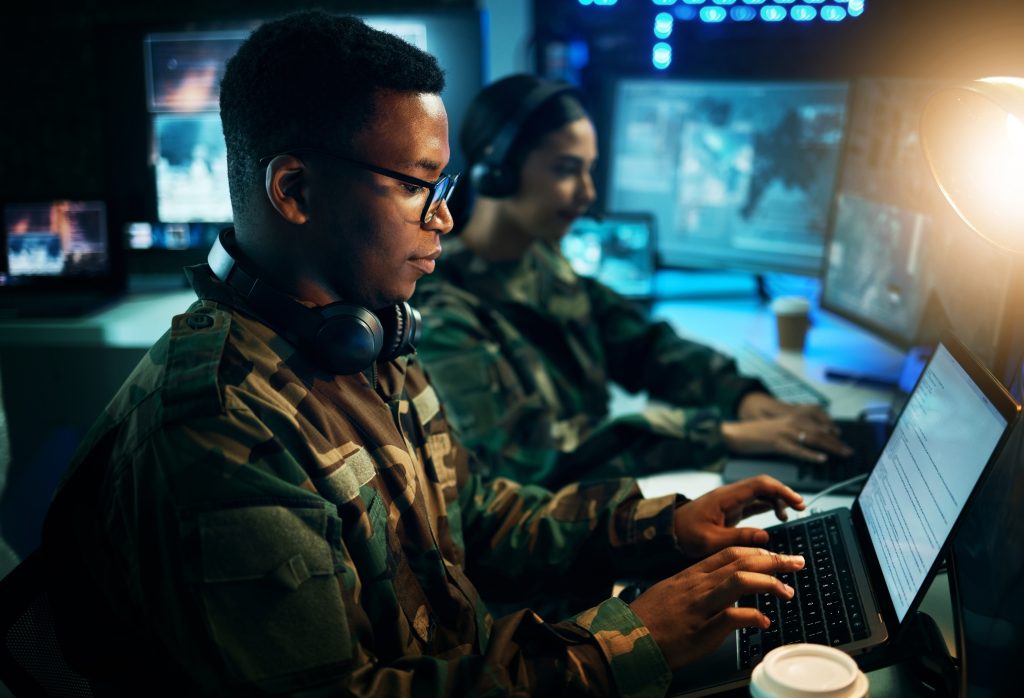 Two people in military uniforms work on laptops in a dimly lit room with multiple monitors displaying digital data and maps.