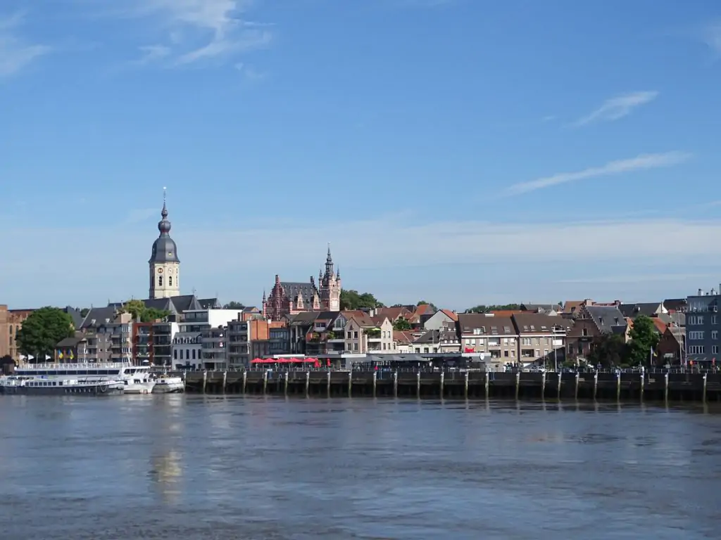 Riverside view of a European town with various buildings, including a church with a tall spire, and a boat docked by the water under a clear blue sky.