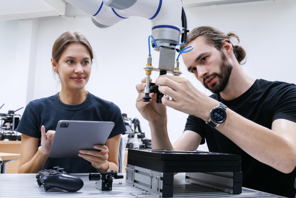 Two people work with a robotic arm; one adjusts a component while the other observes and takes notes on a tablet in a lab setting.