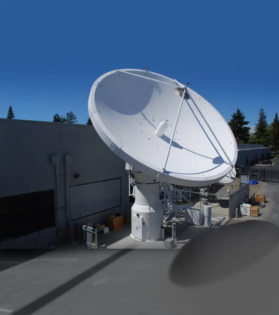 A large white satellite dish antenna is mounted on a platform outside a building under a clear blue sky.