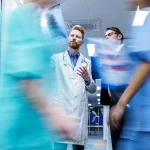 A doctor in a white coat stands in a hospital hallway, while blurred figures in scrubs move quickly past him, suggesting a busy medical environment.