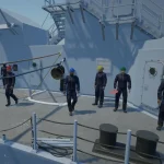 Five people in protective uniforms and colored helmets stand and walk on the deck of a naval ship under clear daylight.