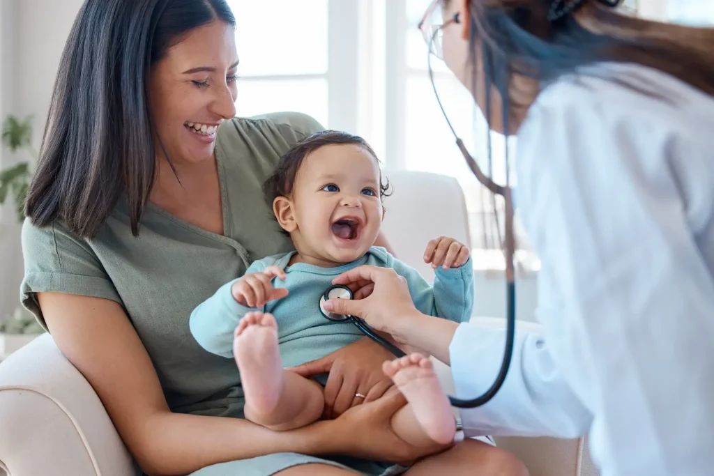 A doctor uses a stethoscope to examine a smiling baby sitting on a woman's lap, both of whom appear happy and relaxed.