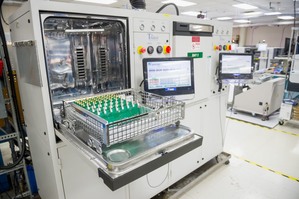 A circuit board is placed in a metal basket inside an industrial machine in a clean manufacturing facility; monitors display system data.
