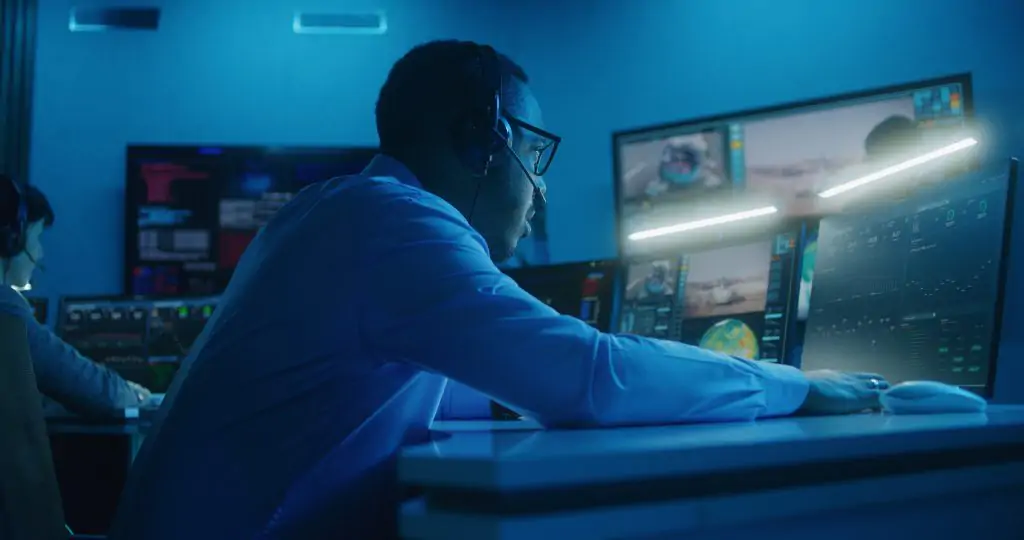 A person wearing a headset sits at a desk with multiple monitors displaying data and images in a dimly lit control room.