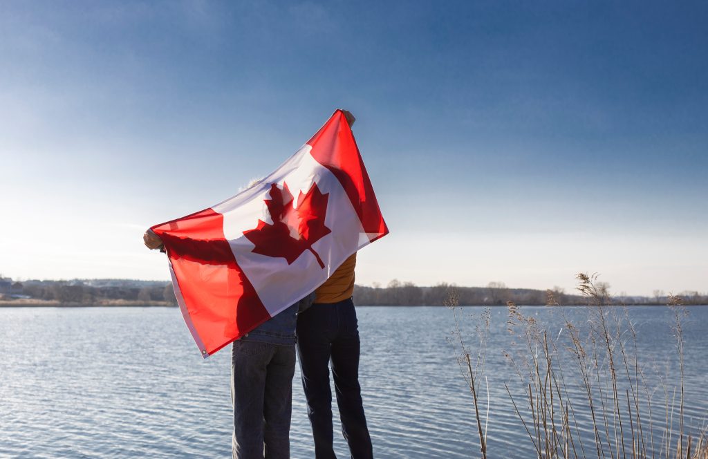 Two people stand by a body of water holding a large Canadian flag, with blue sky and reeds visible in the background.
