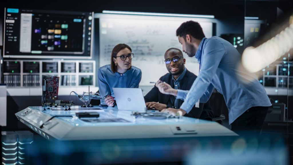Three engineers collaborate at a desk with a laptop and electronic equipment in a high-tech lab, with computer monitors and servers in the background.
