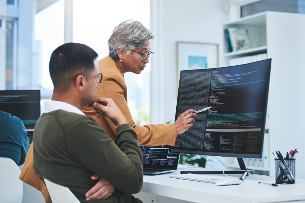 An older woman points at code on a computer screen while instructing a younger man, both seated in a modern office setting with laptops and monitors.