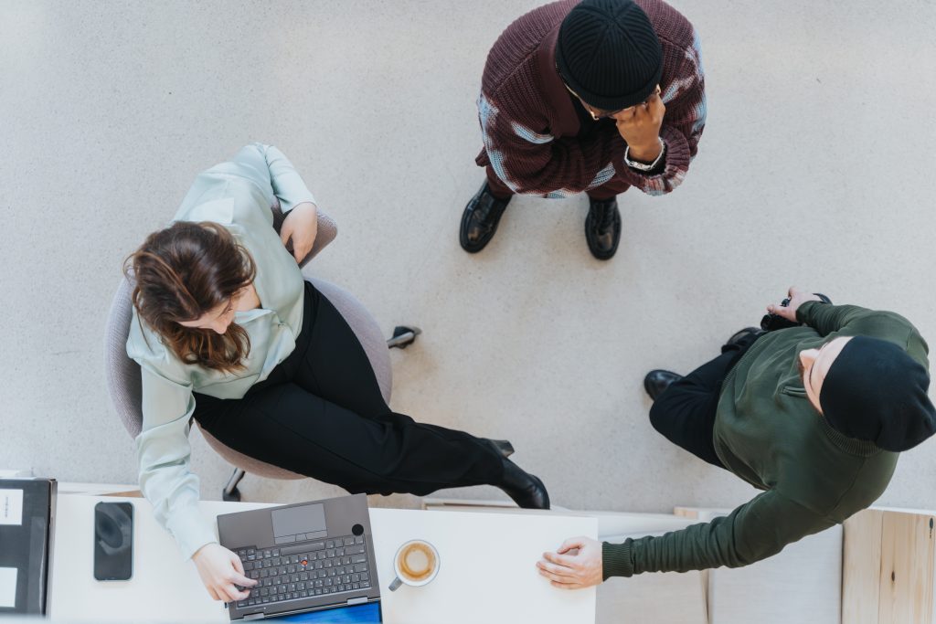 A woman sits at a desk using a laptop while two people stand nearby; the scene is viewed from above.