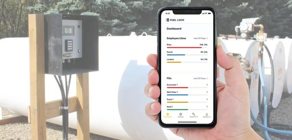 A hand holds a smartphone displaying a fuel management dashboard in front of large outdoor fuel tanks and a control panel.