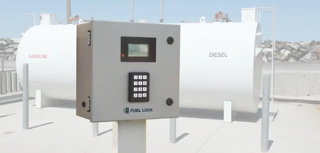 A gray fuel control panel with a keypad and display stands in front of labeled gasoline and diesel storage tanks outdoors.