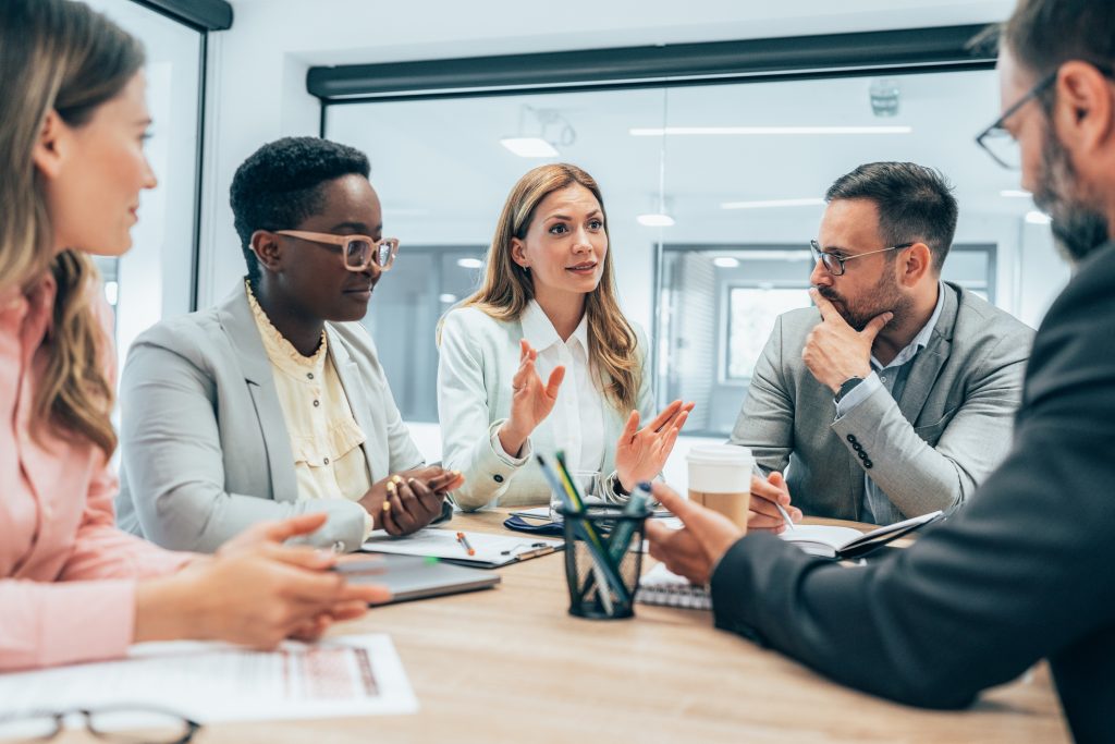 Five people in business attire sit around a conference table, engaged in discussion, with documents, notebooks, and a coffee cup on the table.