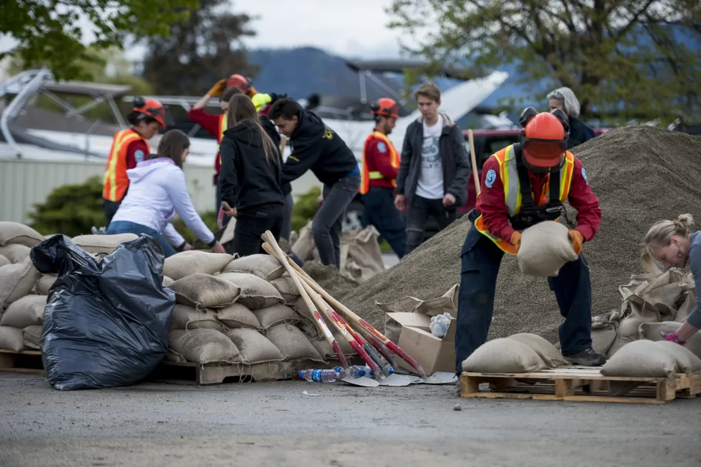 A group of people fill and stack sandbags outdoors, wearing safety gear and working together near a large pile of sand.