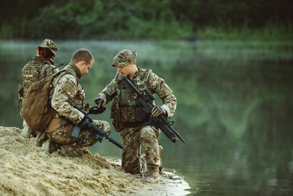 Three soldiers in camouflage uniforms, armed and wearing tactical gear, kneel and interact near the edge of a calm body of water with greenery in the background.