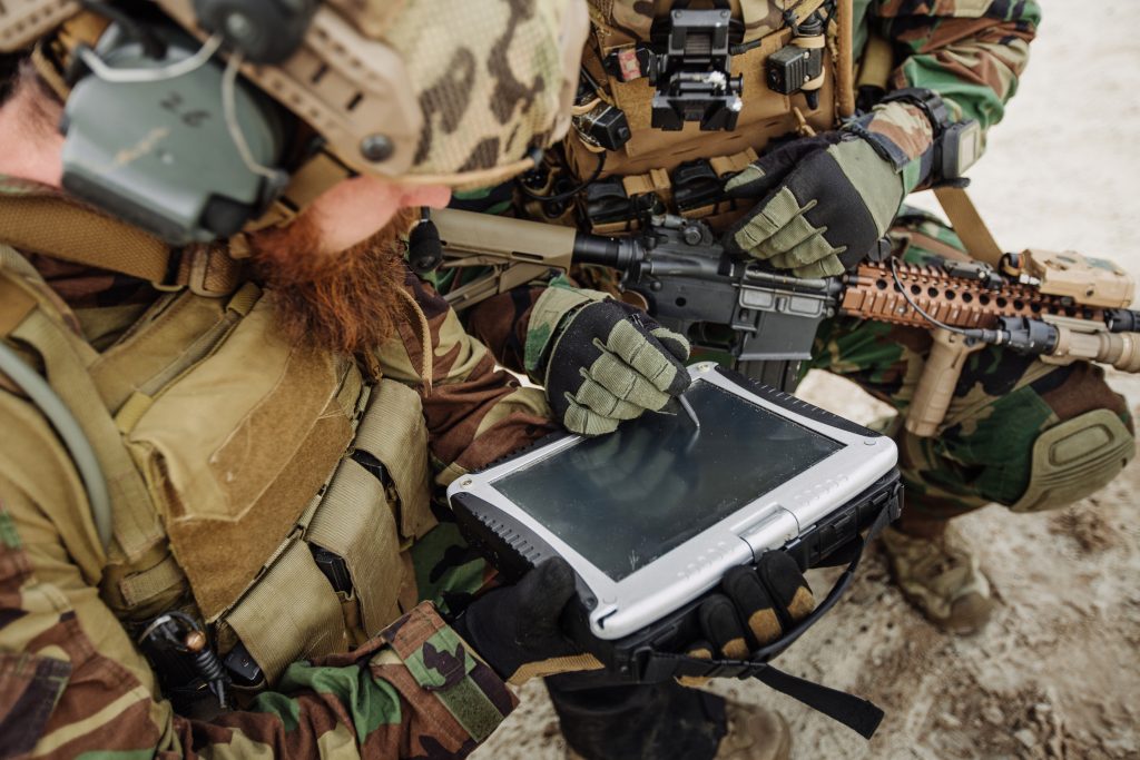 Two soldiers in camouflage uniforms use a rugged tablet device outdoors, with one soldier operating the touchscreen and the other holding a rifle.