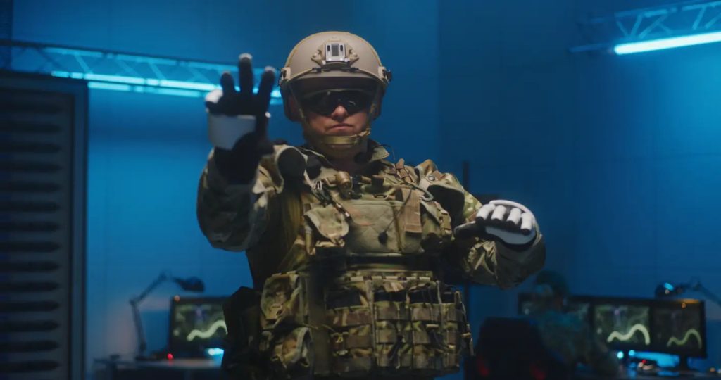A soldier in tactical gear stands indoors with one hand raised, wearing a helmet and goggles, with computer monitors visible in the background.