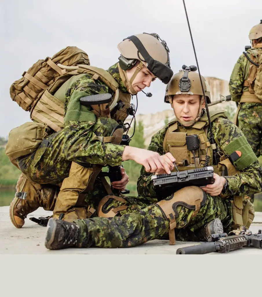 Two soldiers in camouflage uniforms and helmets operate a rugged tablet device outdoors, with one kneeling and the other sitting on the ground. Military gear and equipment are visible.