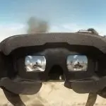 Close-up view of virtual reality goggles with reflections of a desert landscape and a person in the lenses, under a clear blue sky.
