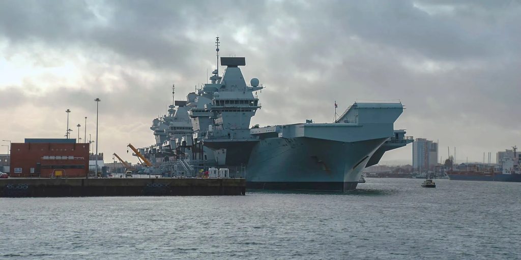A large gray aircraft carrier docked at a port with cranes, buildings, and cloudy skies in the background.