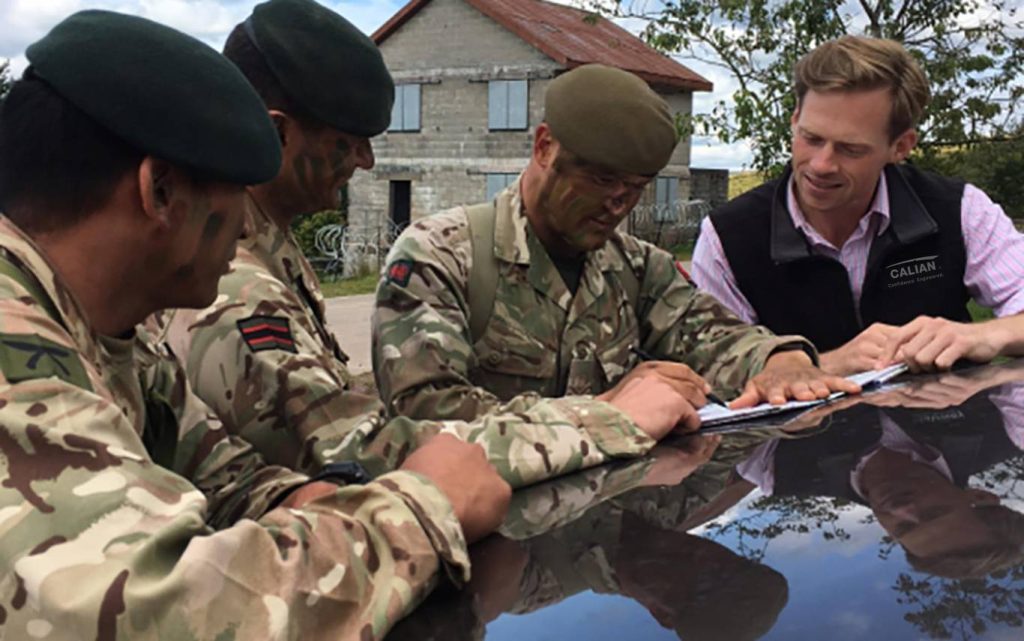 Three soldiers in camouflage uniforms and berets confer with a man in civilian clothing over documents on a car hood, with a building in the background.